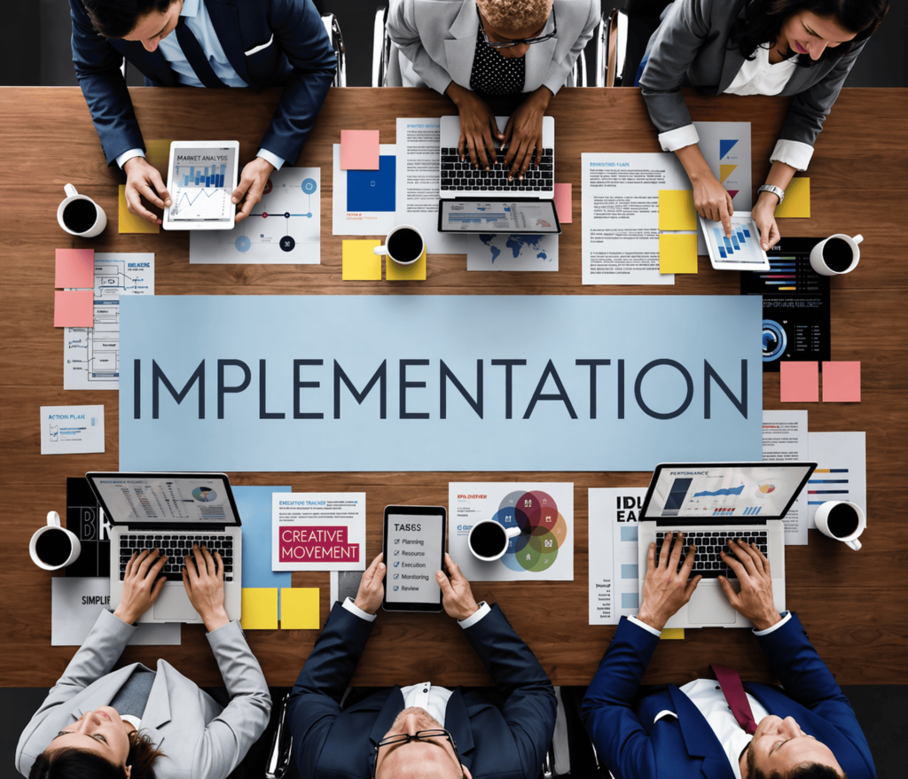 Vista aérea de un equipo de trabajo alrededor de una mesa de madera con portátiles y cafés. En el centro destaca un cartel con la palabra "IMPLEMENTATION" rodeado de notas adhesivas y gráficos de planificación.